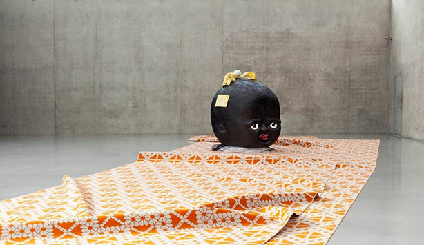 The giant disembodied head of a cartoonish black baby on a orange carpet stares out at the viewer