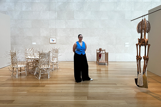 woman in blue shirt and black pants standin in gallery next to sculptures by artist Hugh Hayden