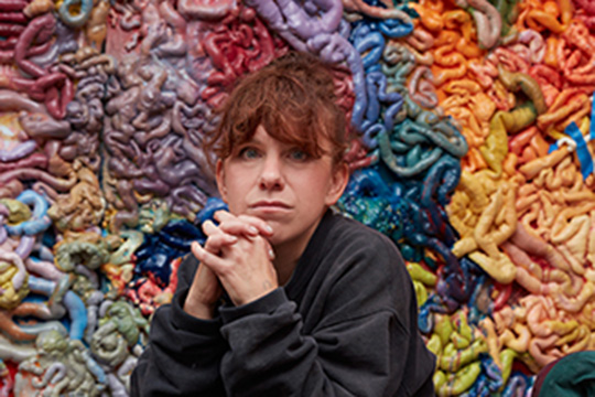 A woman sits in front of a sculpture of colorful intestines. 