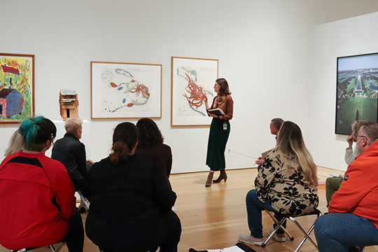 A woman speaks to a group in front of a paintin. 